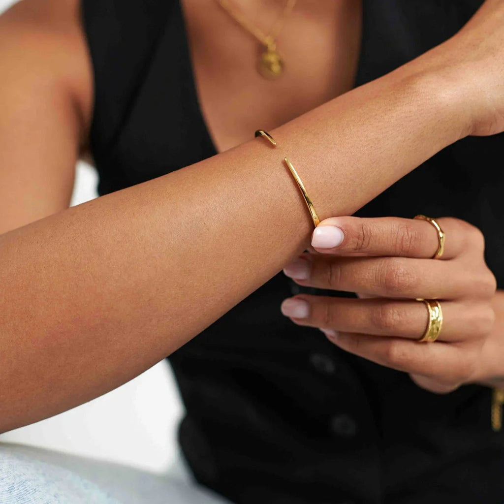 Close-up of a woman's arm wearing a sleek gold cuff bracelet and gold rings, with a black top background