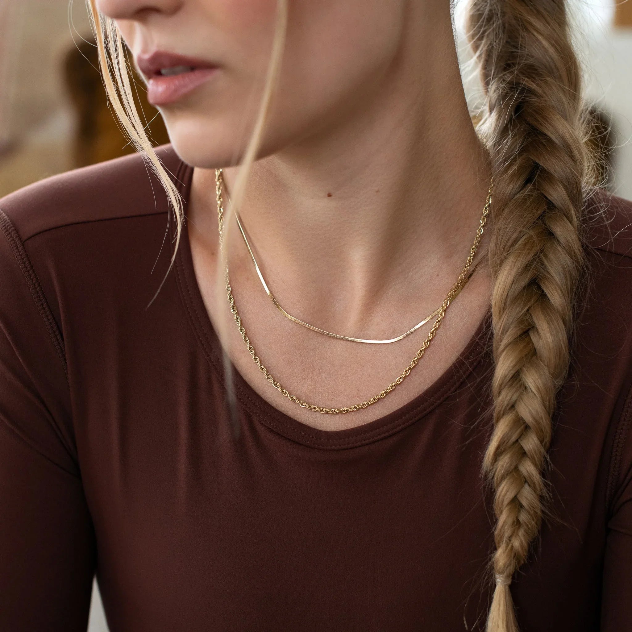 Close-up of a woman with blonde braided hair wearing layered gold necklaces and a brown top