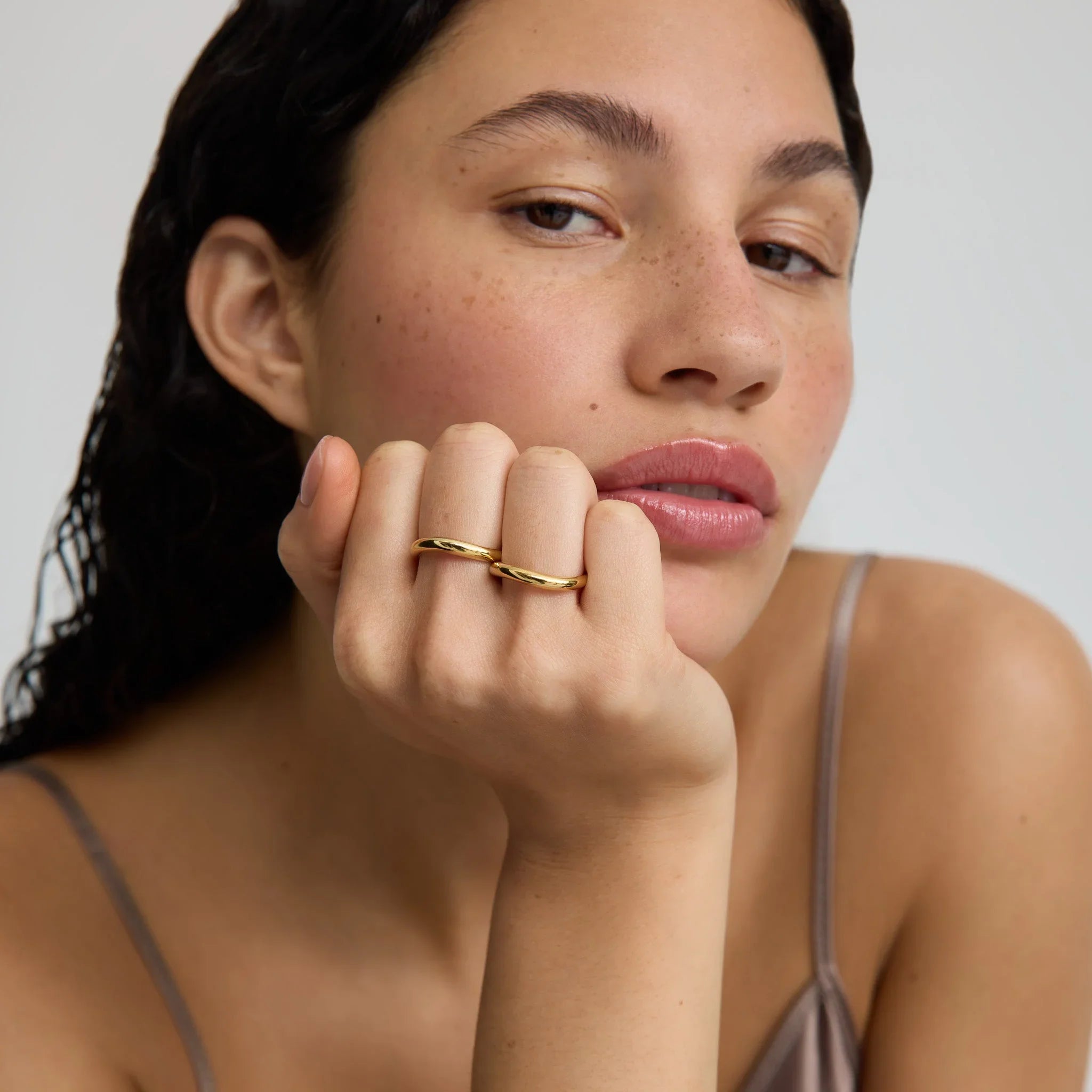 Close-up of woman with natural skin wearing two simple gold rings and a neutral strap top