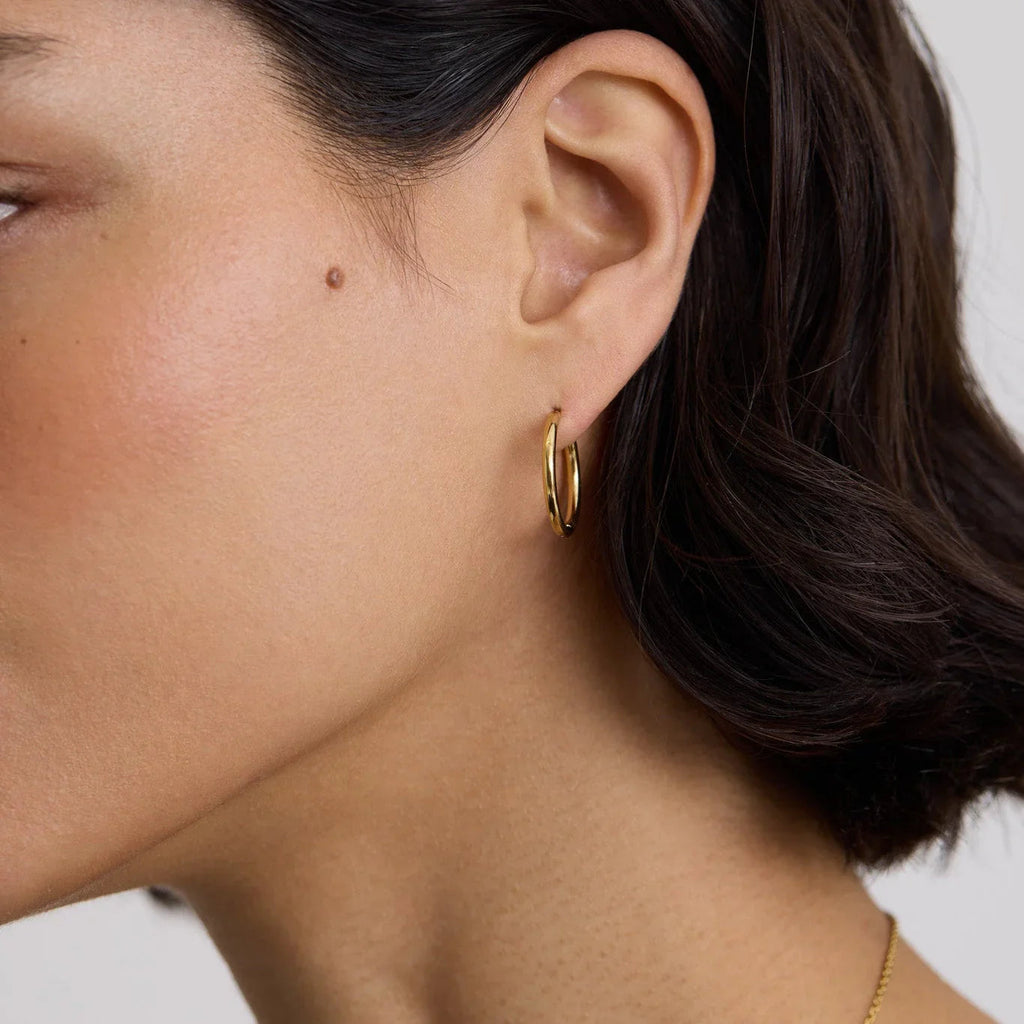 Close-up of a woman's ear wearing a small, polished gold hoop earring with short dark hair